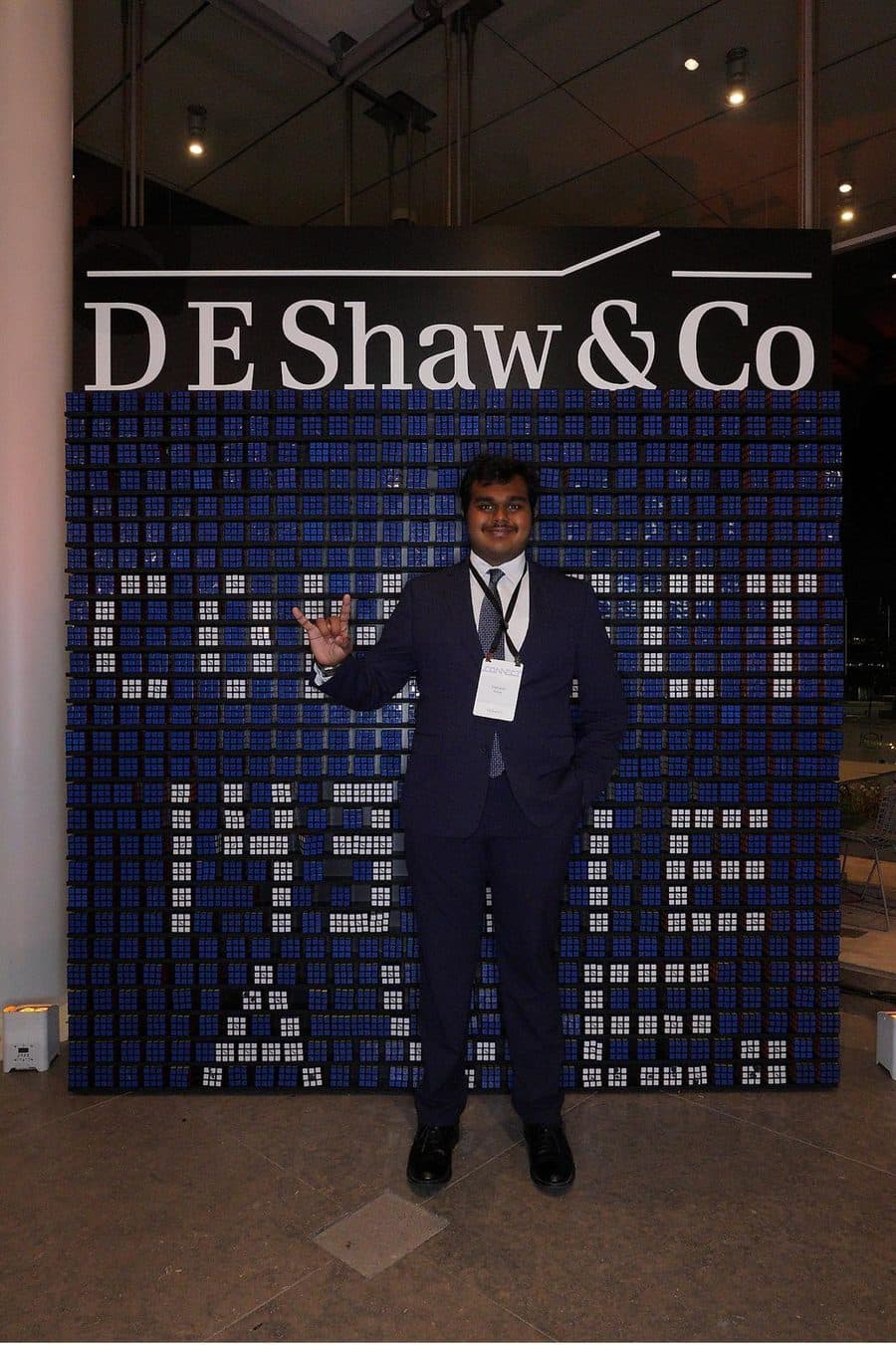 Vishanth in a suit in front of a D. E. Shaw wall made of Rubik cubes